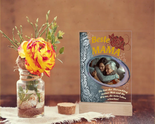 fotoaufsteller-15x20cm-aus-acryl-mit-echtholzsockel-mit-personalisierbaren-foto-silbernen-akzent-auf-der-linken-seite-und-einem-goldgelben-lebensbaum-über-dem-foto-steht-beste-mama-unter-dem-foto-steht-einer-der-fünf-wählbaren-sprüche-der-aufsteller-steht-auf-einem-holztisch-daneben-eine-rose