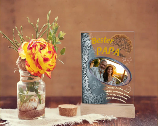 fotoaufsteller-aus-acryl-mit-echtholzsockel-mit-personalisierbaren-foto-silbernen-akzent-auf-der-linken-seite-und-einem-goldgelben-lebensbaum-über-dem-foto-steht-bester-papa-unter-dem-foto-steht-einer-der-fünf-wählbaren-sprüche-der-aufsteller-steht-auf-einem-holztisch-daneben-eine-rose
