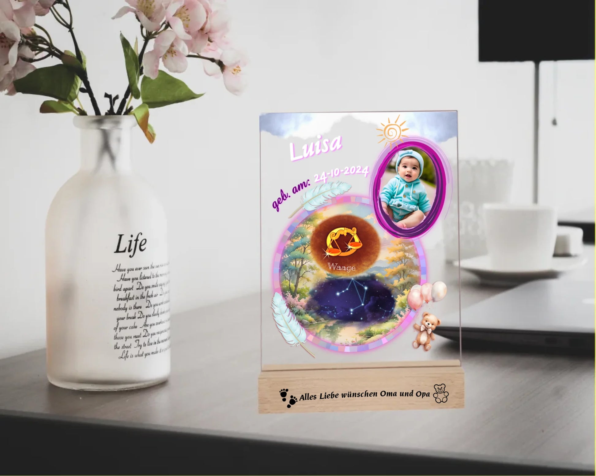 personalisiertes-acrylglas-15x20cm-mit-graviertem-holzsockel-acrylglas-mit-foto-sternzeichen-sternbild-und-name-des-babys-design-in-pastelfarben-überwiegend-in-pink-holzsockel-ist-mit-wunschtext-graviert-fotoaufsteller-steht-auf-einem-schreibtisch-neben-einer-vase-mit-blumen