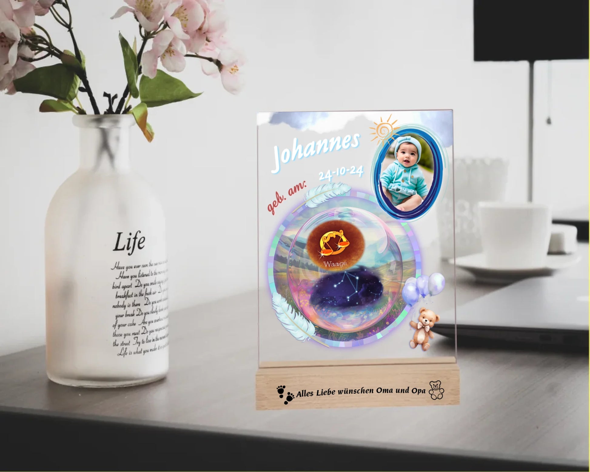 personalisiertes-acrylglas-15x20cm-mit-graviertem-holzsockel-acrylglas-mit-foto-sternzeichen-sternbild-und-name-des-babys-design-in-pastelfarben-überwiegend-in-blau-holzsockel-ist-mit-wunschtext-graviert-fotoaufsteller-steht-auf-einem-schreibtisch-neben-vase-mit-blumen