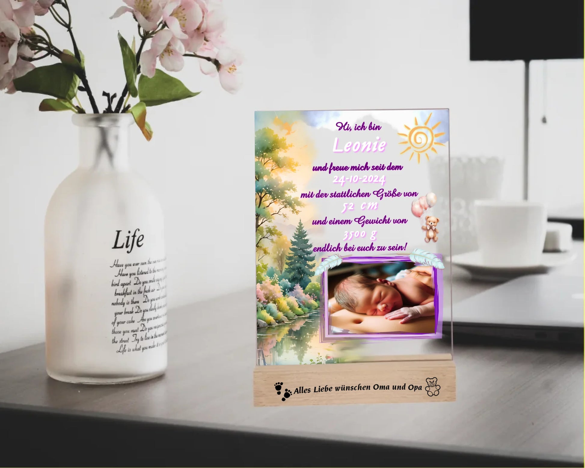 acrylglas-fotoaufsteller-15x20cm-mit-graviertem-standfuß-aus-holz-personalisierter-steckbrief-mit-geburtsdaten-und-foto-hauptfarbe-des-designs-pink-und-pasteltoene-standfuss-mit-wunschtext-graviert-fotoauftseller-steht-auf-schreibtisch-neben-einer-vase-mit-blumen