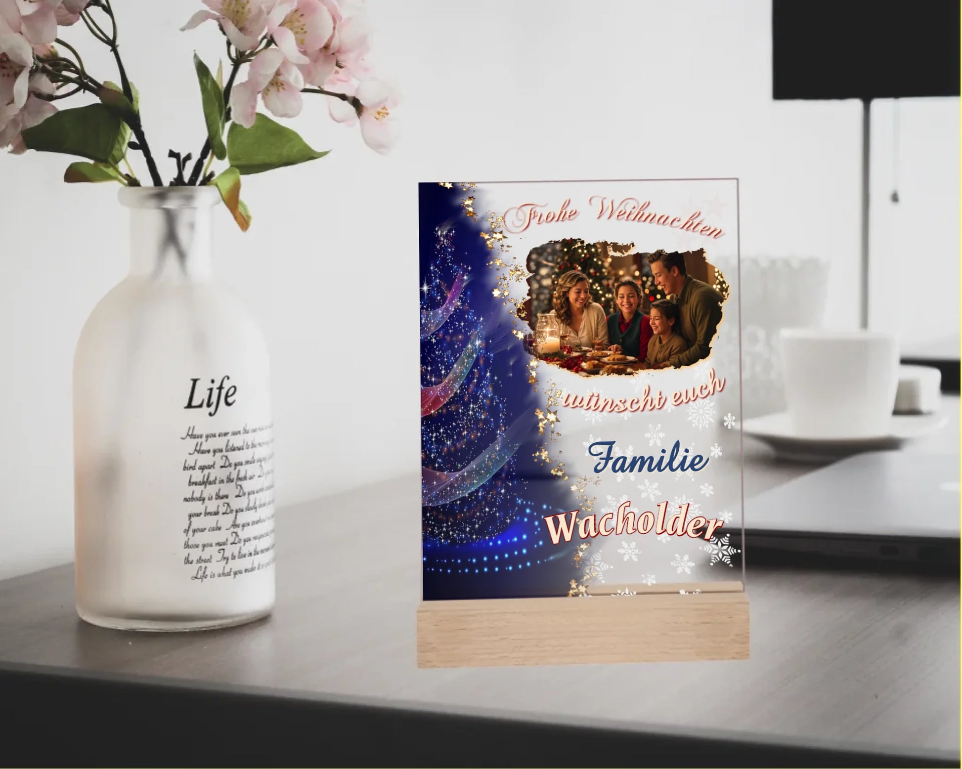 Mockacrylglas-15x20cm-mit-standfuß-aus-echtholz-personalisiert-mit-familienfoto-und-familienname-in -der-linken-haelfte-ist-ein-weihnachtsbaum-mit-bunten-lichtern-und-dunkelblauem-hintergrund-fließender-uebergang-zur-transparentnen-hälfte-mit-foto-name-und-schriftzug-frohe-weihnachten-wuenscht-euch-familie-name-der-familie-fotoaufsteller-steht-auf-einem-schreibtisch-neben-einer-vase-mit-blumen