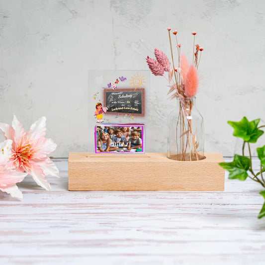 fotoaufsteller-aus-acrylglas-8x11,5cm-personalisiert-mit-foto-und-name-der-schule-mit-kleiner-vase-mit-trockenblumen-auf-einem-hellen-echtholz-sockel-fotoaufsteller-steht-neben-einer-blume