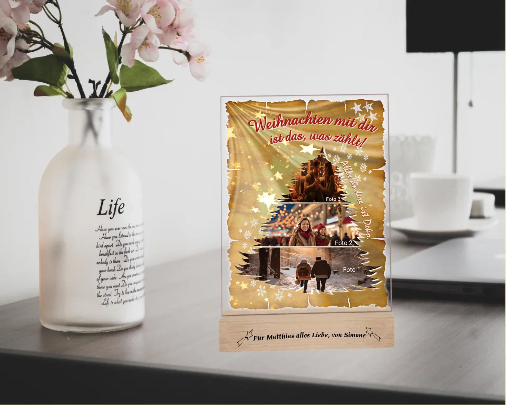fotoaufsteller-acryl-15x20cm-mit-graviertem-standfuss-acrylglas-mit-3-teiliger-foto-collage-in-tannenbaum-form-darueber-schriftzug-weihnachten-mit-dir-ist-das-was-zaehlt-und-neben-der-collage-steht-alles-andere-ist-deko-mit-einem-hintergrund-optik-goldenes-papier-mit-sternen-und-schneeflocken-fotoaufsteller-steht-auf-schreibtisch-neben-einer-vase-mit-blumen