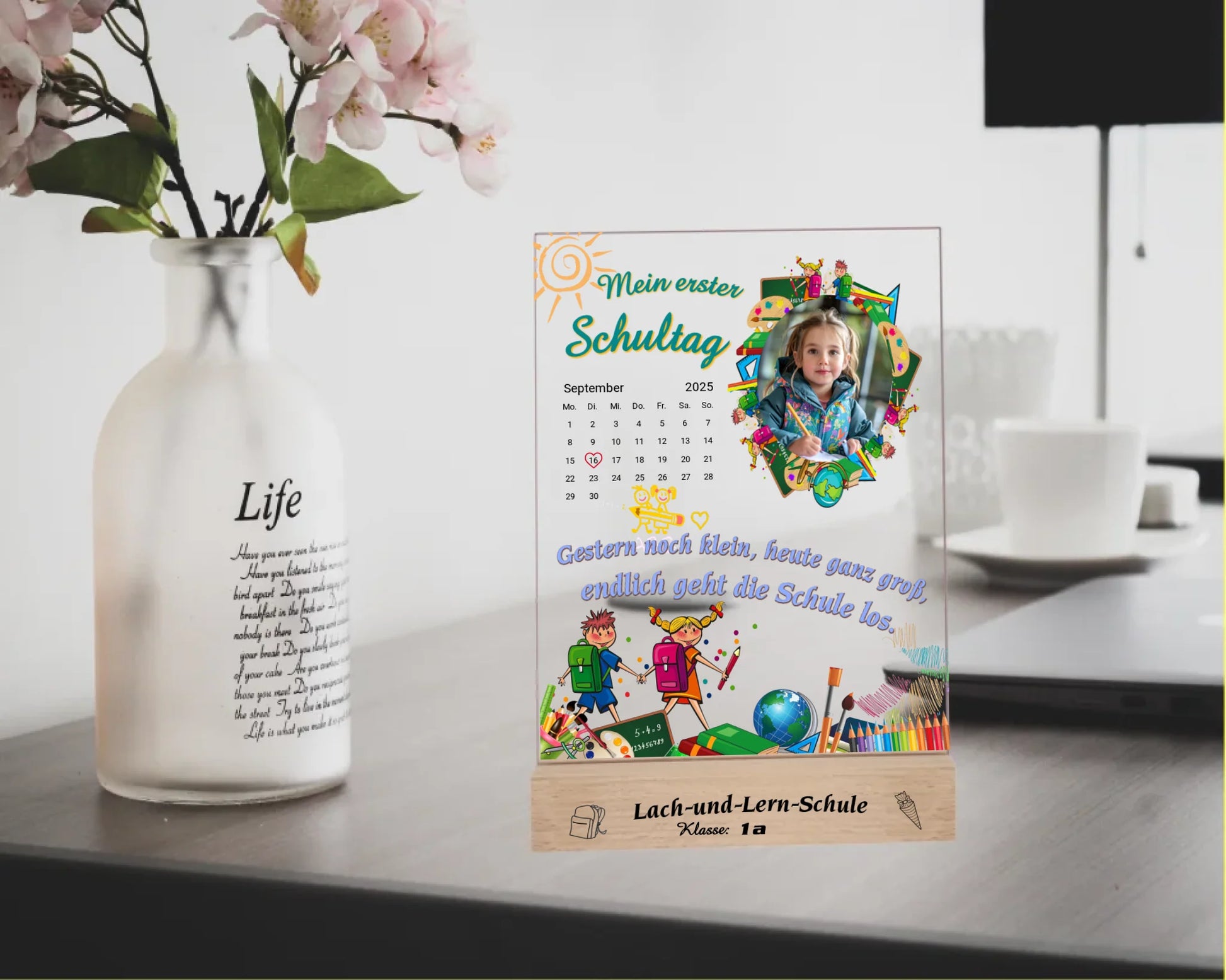 personalisierter-fotoaufsteller-aus-acryl-15x20cm-mit-foto-und-kalenderblatt-zum auswählen-des-datums-zwischen-bunten-schulmotiven-auf-graviertem-holzsockel-mit-name-der-schule-und-der-klasse-fotoaufsteller-steht-auf-schreibtsch