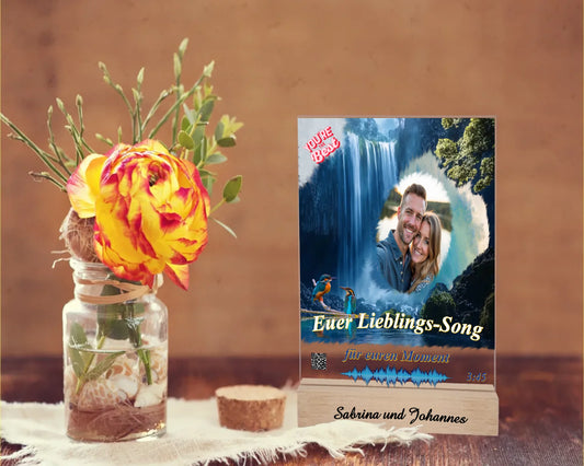 acrylaufsteller-15x20cm-personalisiert-mit-foto-titel-und-interpret-des-ausgewählten-songs-mit-qr-code-und-gravierten-namen-am-holzsockel-design-hintergrund-ein-wasserfall-in-strahlendem-blau-aufsteller-steht-auf-einem-holztisch-neben-einer-rose