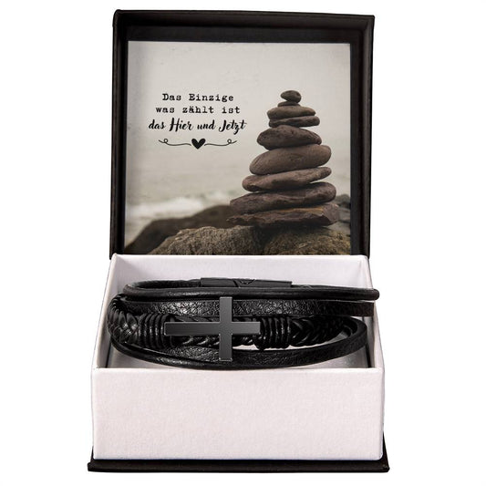 geflochtenes-kunsteder-armband-mit-edelstahl-kreuz-in-schwarzem-finish-in-der-schwarz-weißen-satndarbox-darüber-die-karte-mit-gestapelten-steinen-und-botschaft-das-einzige-was-zählt-ist-das-hier-und-jetzt