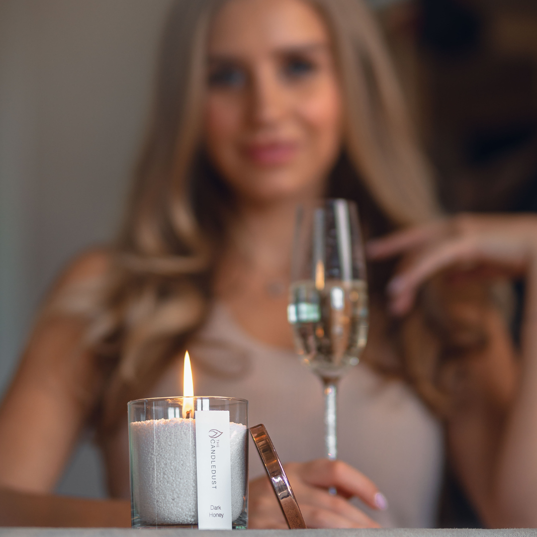 kerze-aus-wachsgranulat-in-transparentem-glas-brennt-im-vordergrund-im-hintergrund-sitzt-eine-blonde-frau-mit-sektglas