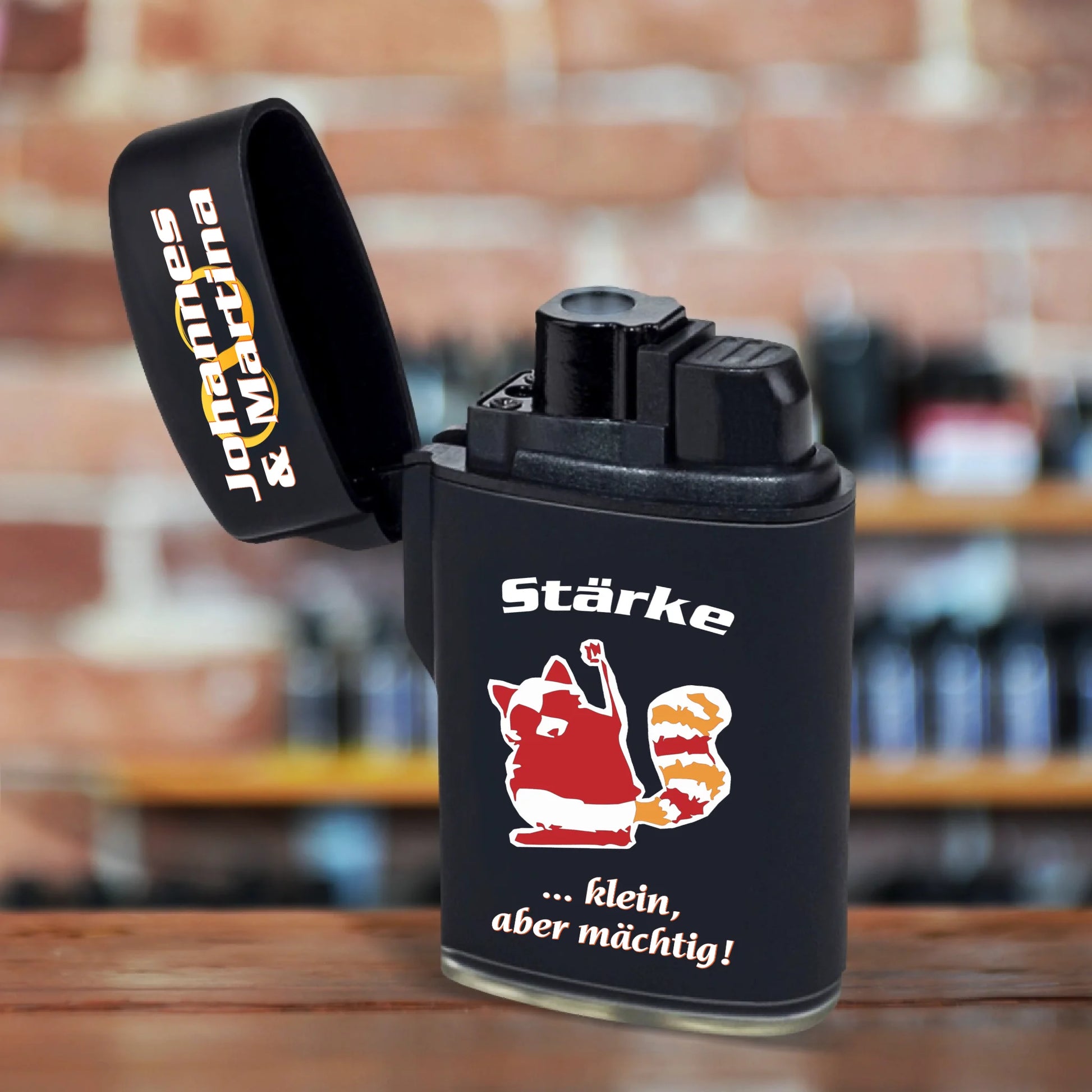 jetflame-feuerzeug-schwarz-mit-deckel-geoeffnet-nikolausgeschenk-kollegen-personalisierbar-mit-namen-und-charaktereigenschaft-dazu-erscheint-ein-passendes-motiv-hier-waschbär-in-rot-orange-und-weißer-text-staerke-klein-aber-maechtig-feuerzeug-steht-auf-tischplatte