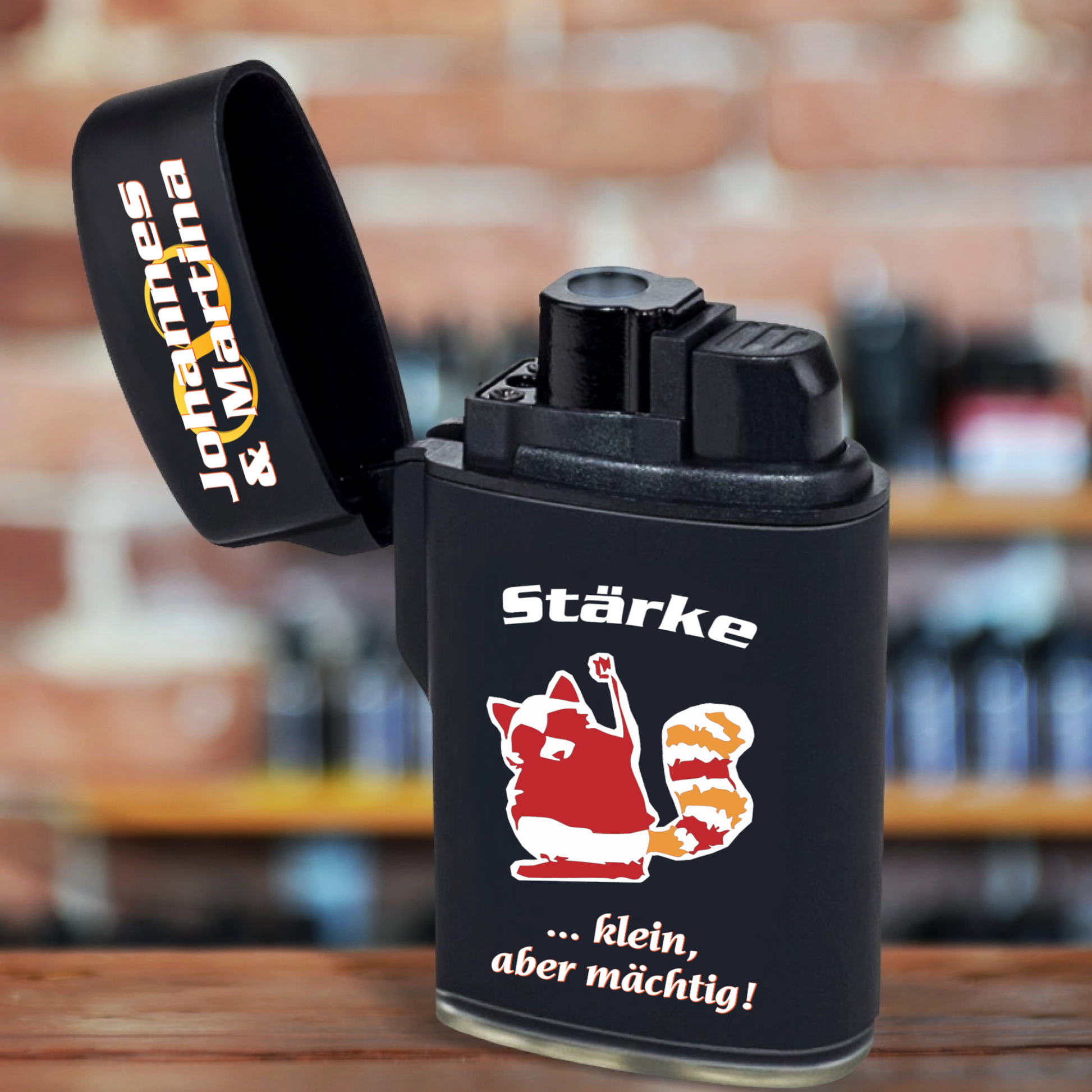 jetflame-feuerzeug-schwarz-mit-deckel-geoeffnet-nikolausgeschenk-kollegen-personalisierbar-mit-namen-und-charaktereigenschaft-dazu-erscheint-ein-passendes-motiv-hier-waschbär-in-rot-orange-und-weißer-text-staerke-klein-aber-maechtig-feuerzeug-steht-auf-tischplatte