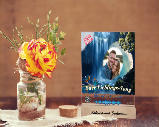 acrylaufsteller-15x20cm-personalisiert-mit-foto-titel-und-interpret-des-ausgewählten-songs-mit-qr-code-und-gravierten-namen-am-holzsockel-design-hintergrund-ein-wasserfall-in-strahlendem-blau-aufsteller-steht-auf-einem-holztisch-neben-einer-rose