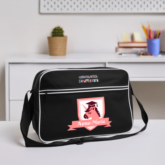 umhaengetasche-40x28cm-schwarz-mit-weissen-kontraststreifen-mit-2-reissverschluessen-und-umhaengegurt-design-pinkes-wappen-mit-einhorn-darnter-pinker-banner-mit-personalisiertem-namen-ganz-oben-auf-der-tasche-schriftzug-kindergarten-ist-durchgestrichen-darunter-steht-in-fetter-schrift-schule-tasche-steht-auf-einem-schreibtisch