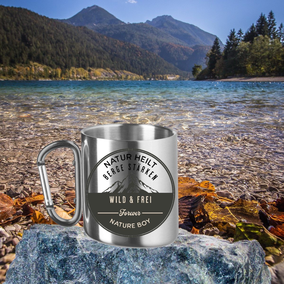 edelstahltasse-mit-karabiner-als-henkel-steht-auf-einem-stein-am-flussufer-im-hintergrund-sind-berge-zu-sehen-auf-der-tasse-ein-bergmotiv-plus-aufschrift-nature-boy-wild-und-frei