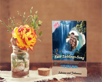 acrylaufsteller-15x20cm-personalisiert-mit-foto-titel-und-interpret-des-ausgewählten-songs-mit-qr-code-und-gravierten-namen-am-holzsockel-design-hintergrund-ein-wasserfall-in-strahlendem-blau-aufsteller-steht-auf-einem-holztisch-neben-einer-rose