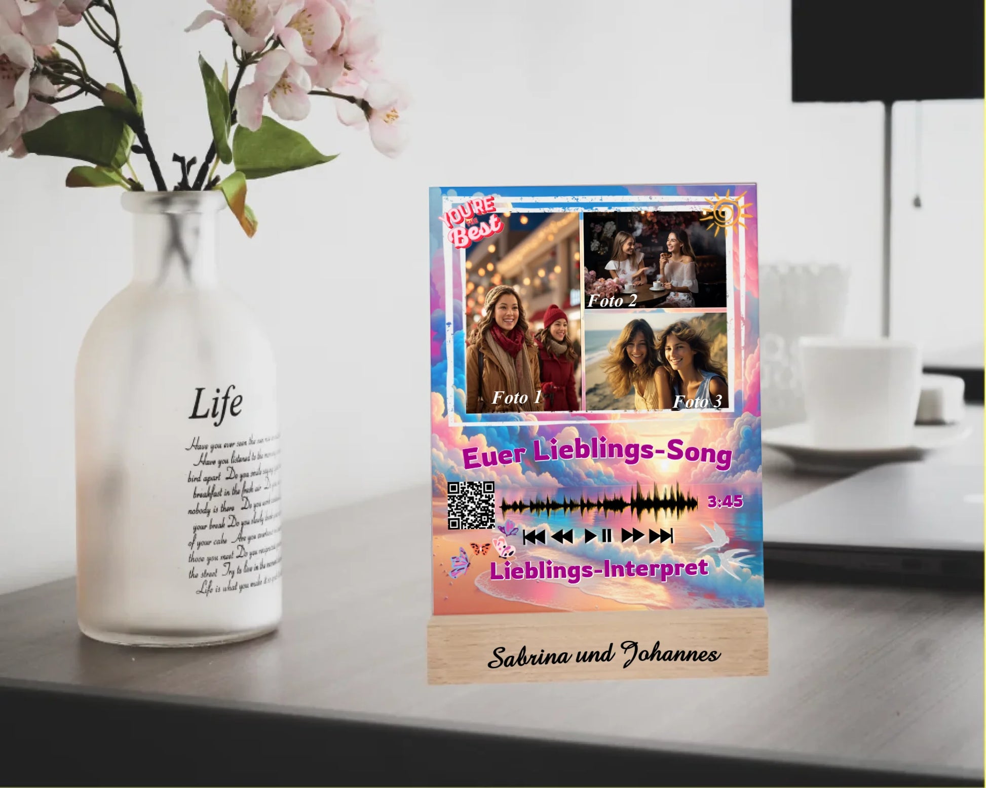 acrylglas-15x20cm-mit-graviertem-standfuss-aus-echtholz-acrylglas-mit-3-fotos-und-ausgewähltem-songtitel-und-interpret-mit-qr-code-design-hintergrund-pastell-rosa-und blau-in-verschiedenen-farbtoenen-standfss-graviert-mit-namen-oder-wunschtext-acrylaufsteller-steht-auf-schreibtisch-neben-vase-mit-blumen