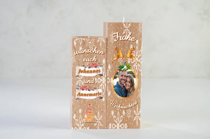 teelichthalter-massiv-holz-2er-set-4,5x4,5cm-12-und-14cm-hoch-personalisiert-mit-foto-und-zwei-namen-weihnachtliches-design-kerzenhalter-rechts-mit-schneeflocken-2-rentier-umrissen-in-gold-ueber-dem-foto-mit-ovalem-goldenem-rahmen-ganz-oben-steht-frohe-und-ganz-unten-steht-weihnachten-design-kerzenhalter-links-schriftzug-wuenschen-euch-dann-die-persnalisierten-namen-auf-schriftrolle-teelichthalter-vor-neutralem-weissen-hintergrund